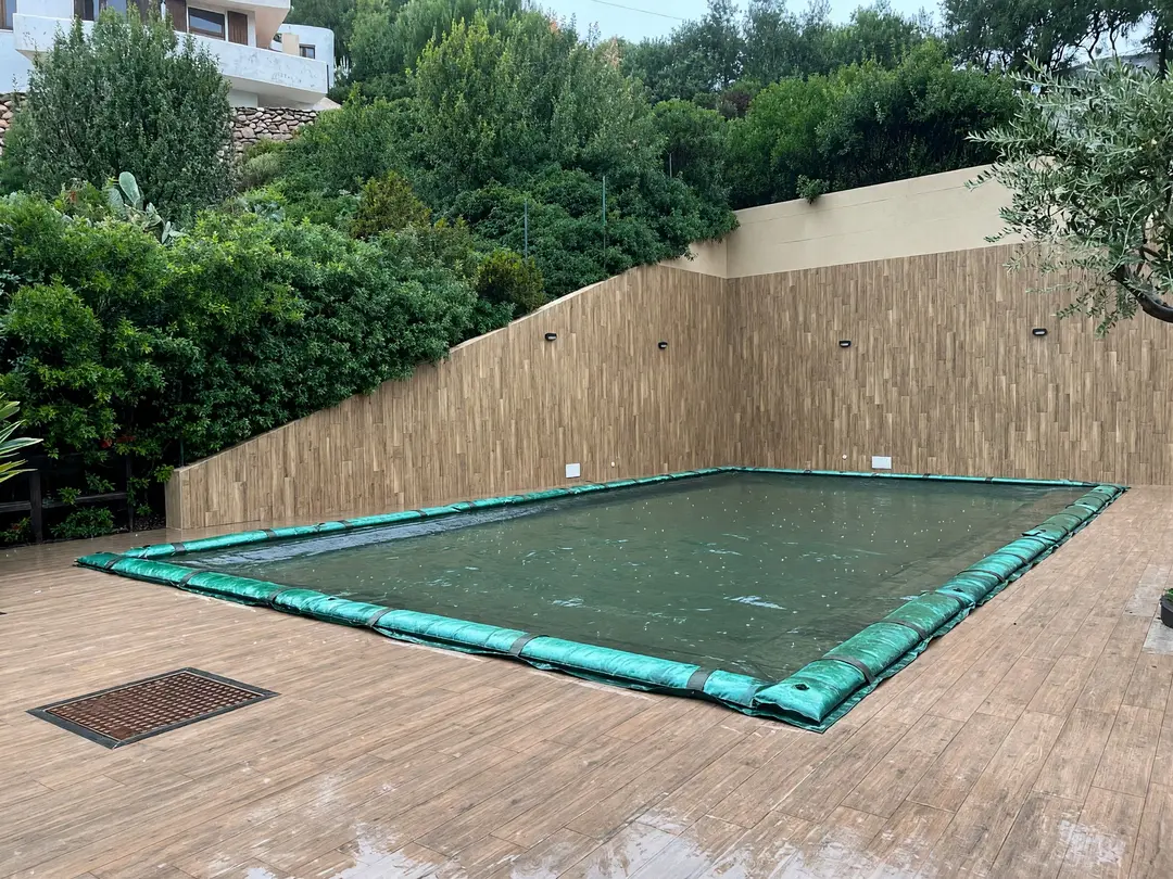 Green winter cover with water tubes on a rectangular pool, between a tall wall and greenery